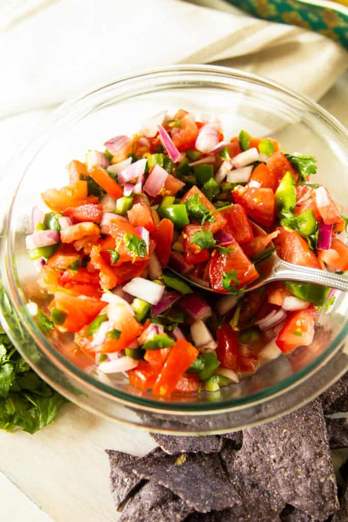 A bowl of pico de gallo with chips and cilantro to its side.