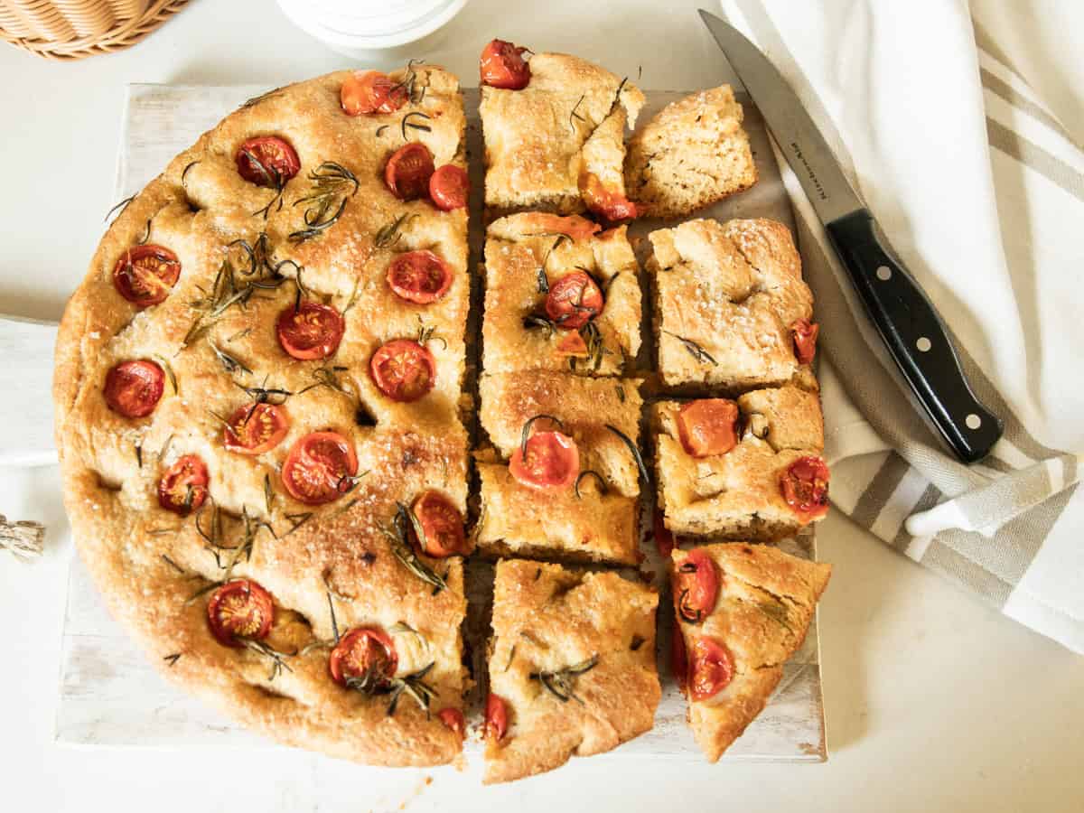 A round focaccia bread on a cutting board with half of it sliced into square pieces.