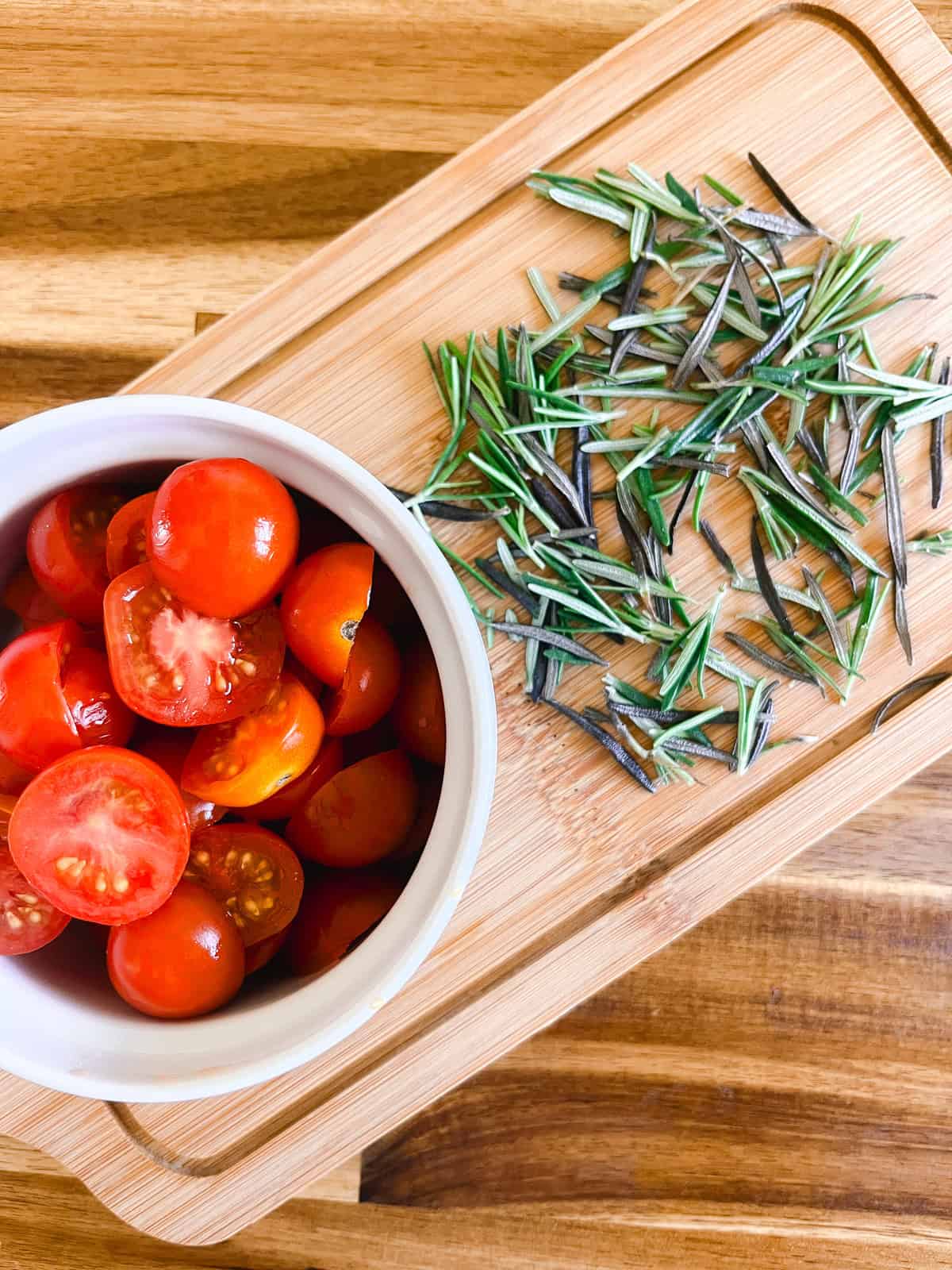 A bowl of halved cherry tomatoes and chopped rosemary on a cutting board.