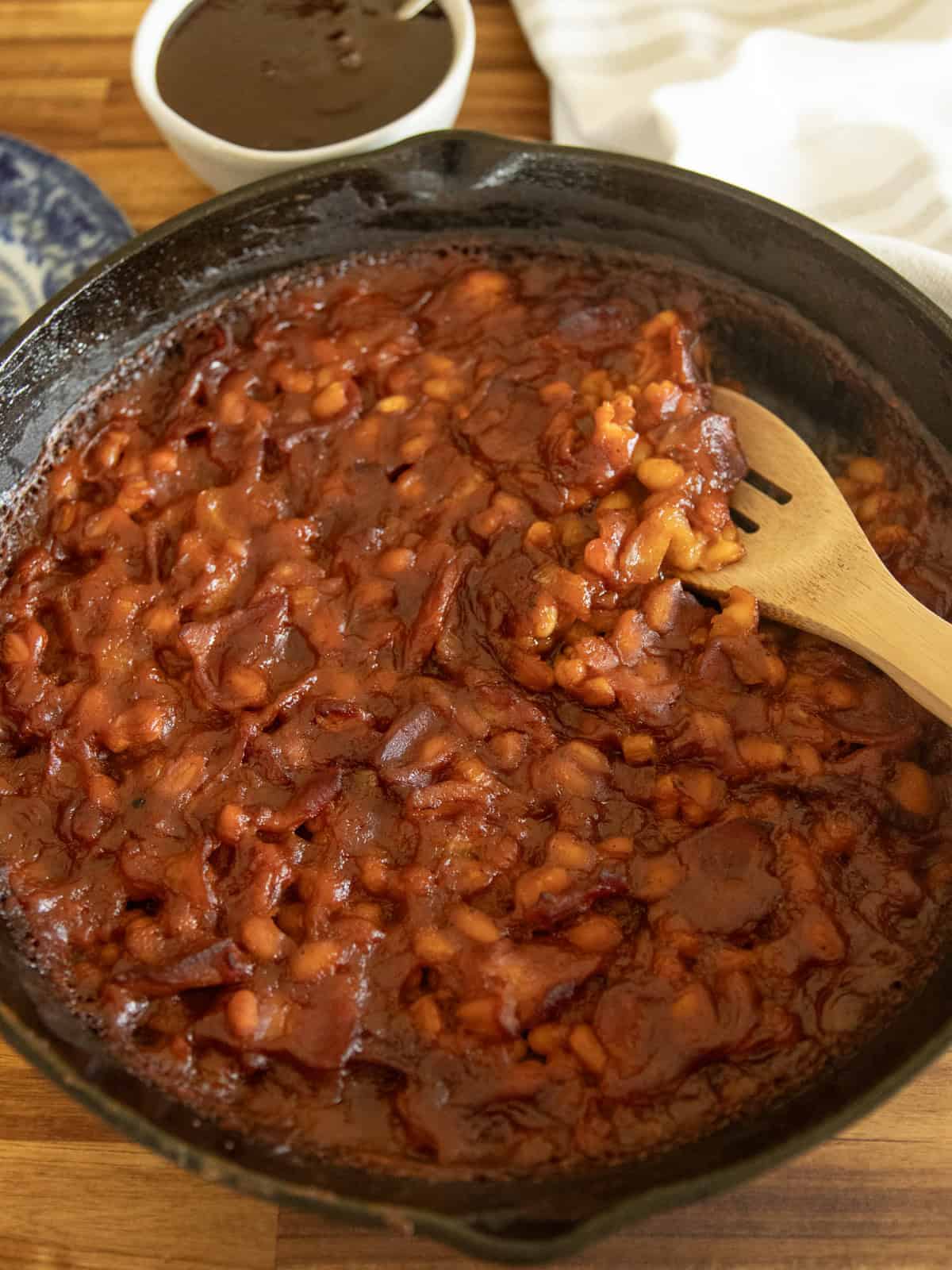 Scooping baked beans out of a cast iron skillet with a wooden spoon.