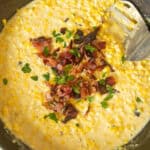 Scooping creamed corn out of a cast iron skillet with bacon on top.