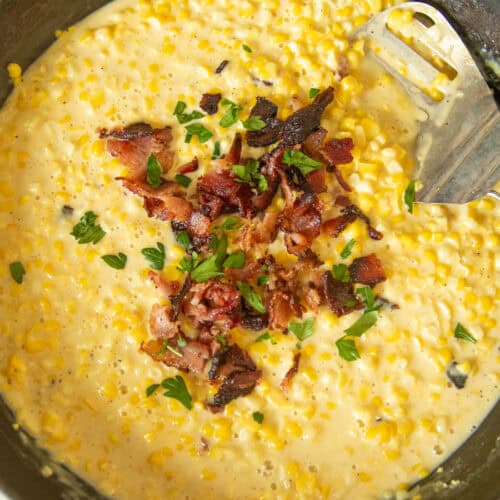 Scooping creamed corn out of a cast iron skillet with bacon on top.