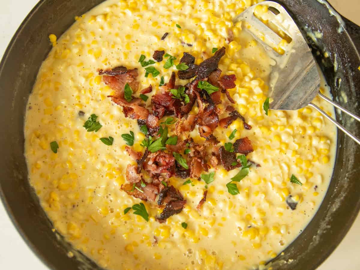 Scooping creamed corn out of a cast iron skillet with bacon on top.