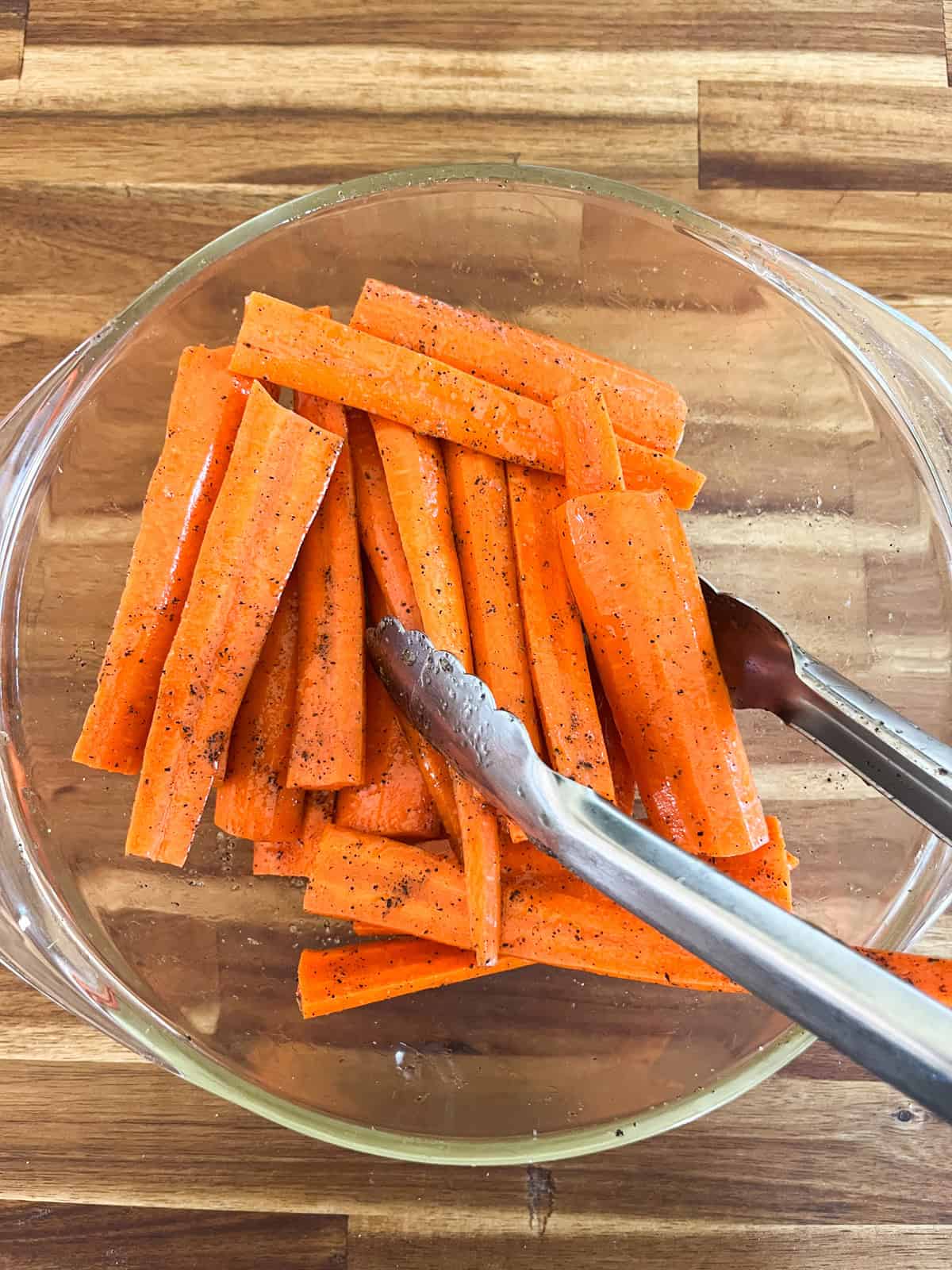 Tossing cut carrots in olive oil in a glass mixing bowl with tongs.