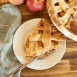 A slice of apple pie with an apple pice, napkin, and whole apples on a wooden counter.