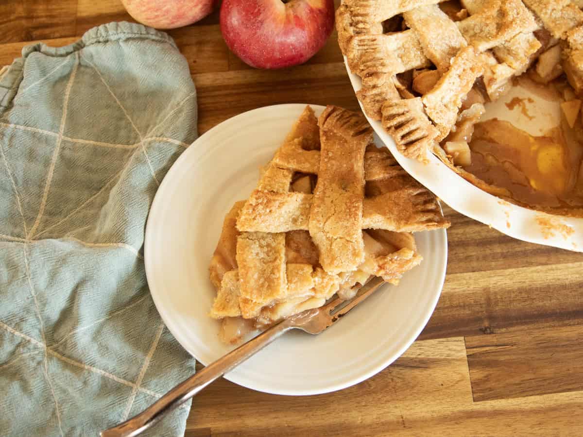 A slice of apple pie with an apple pice, napkin, and whole apples on a wooden counter.
