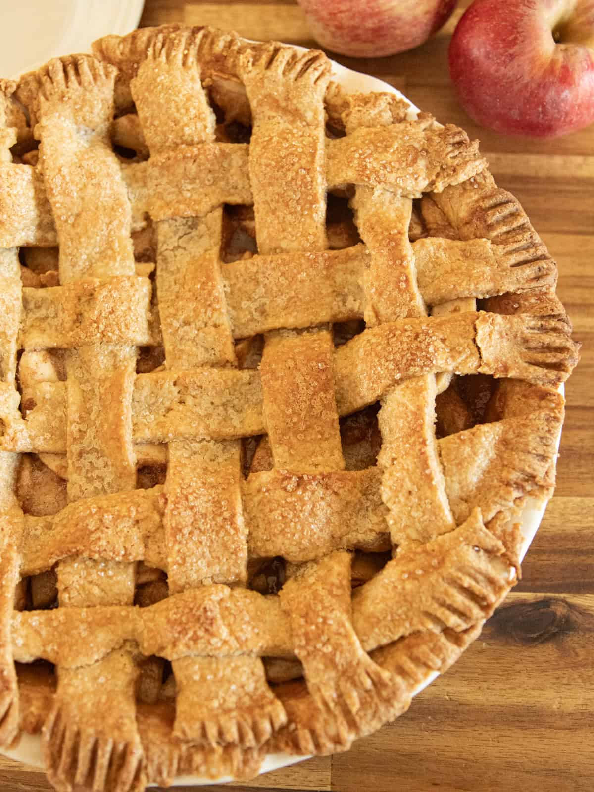 A lattice apple pie next to apples on a wooden backdrop.