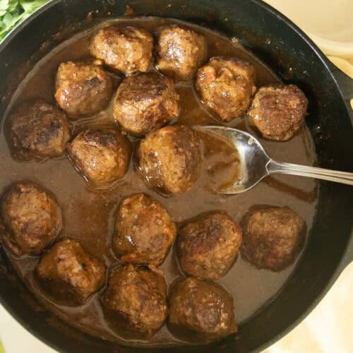 Scooping a meatball with gravy out of a cast iron skillet.
