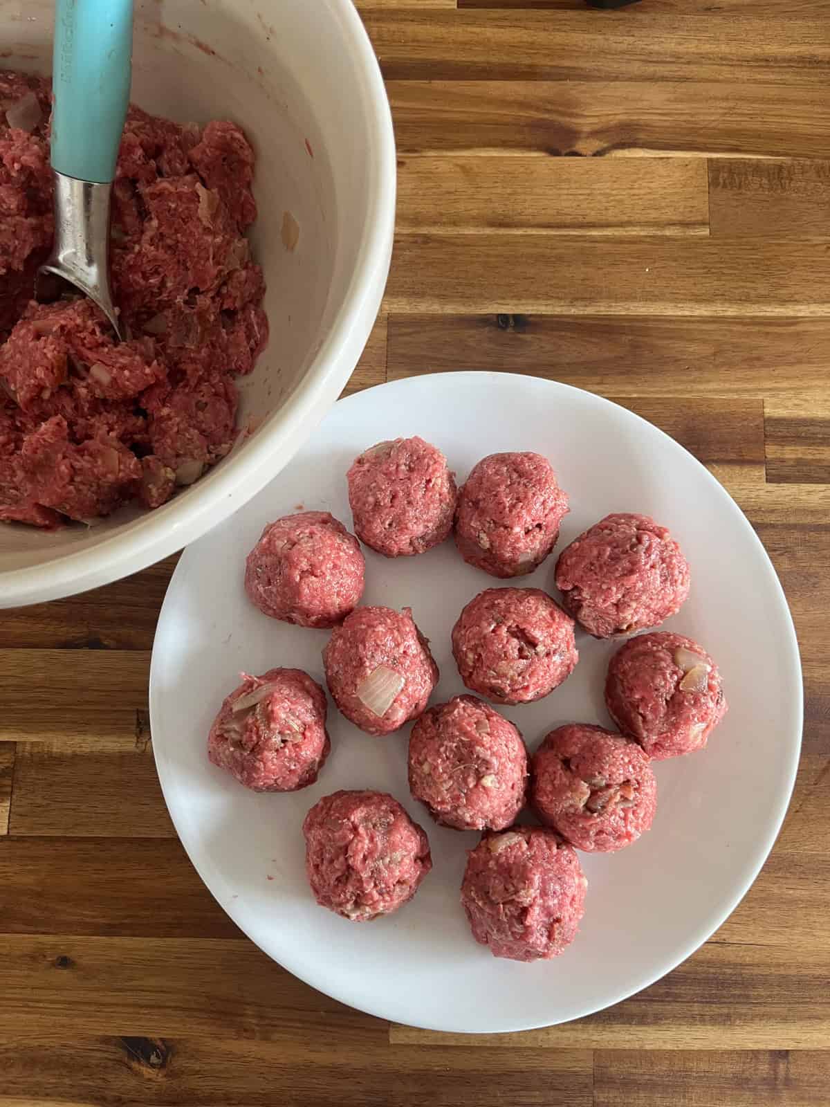 Uncooked meatballs shaped on a white plate with a bowl of meatball mix next to it.