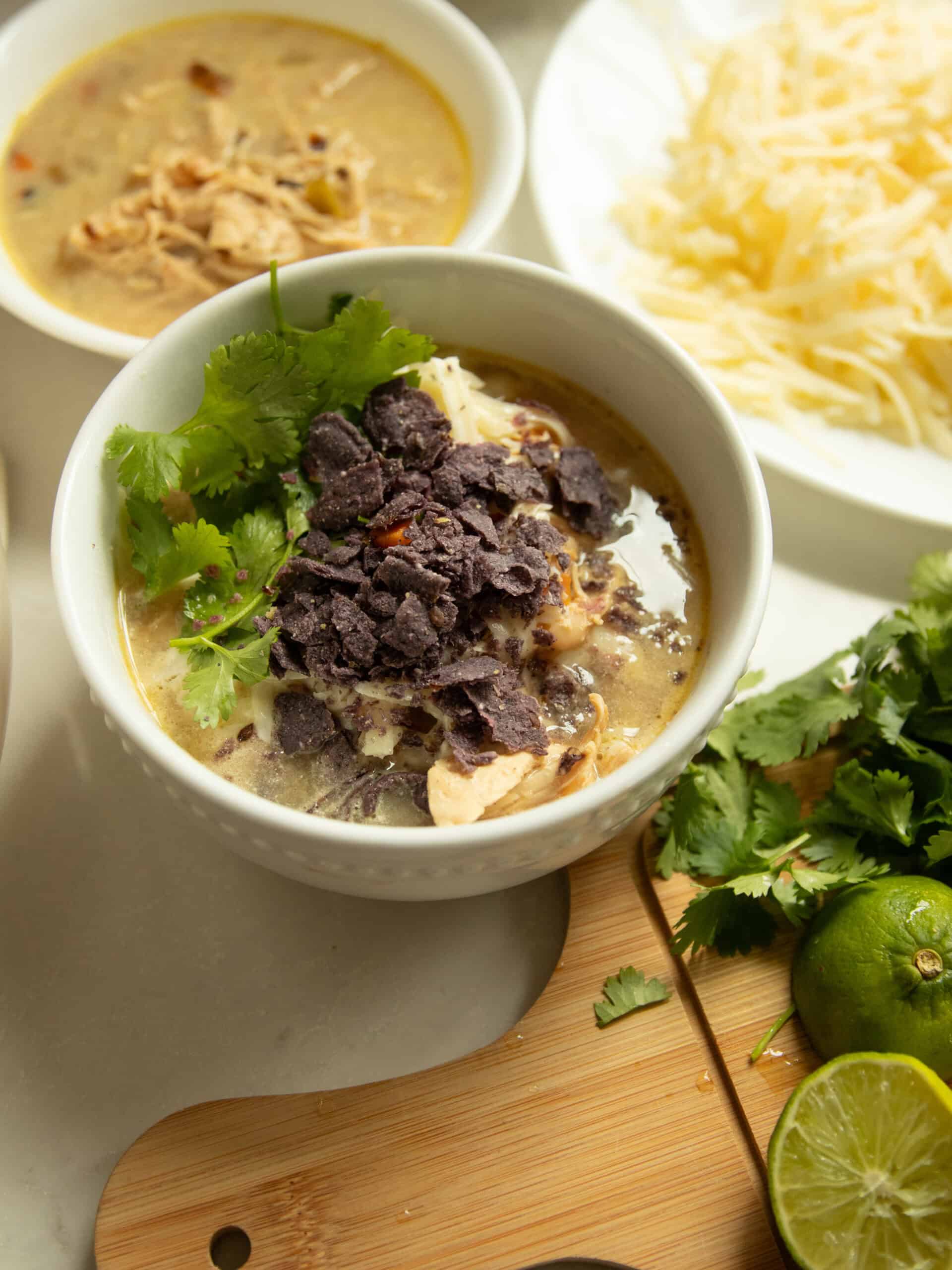 A bowl of white chicken chili with toppings, a bowl of chili without toppings, and toppings surrounding bowl bowls.